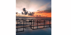 Sunset view from the infinity pool deck at Coral Rock by Bansei in Hikkaduwa, featuring a dark wooden railing reflecting across calm water against a vibrant orange and purple twilight sky over the Indian Ocean.